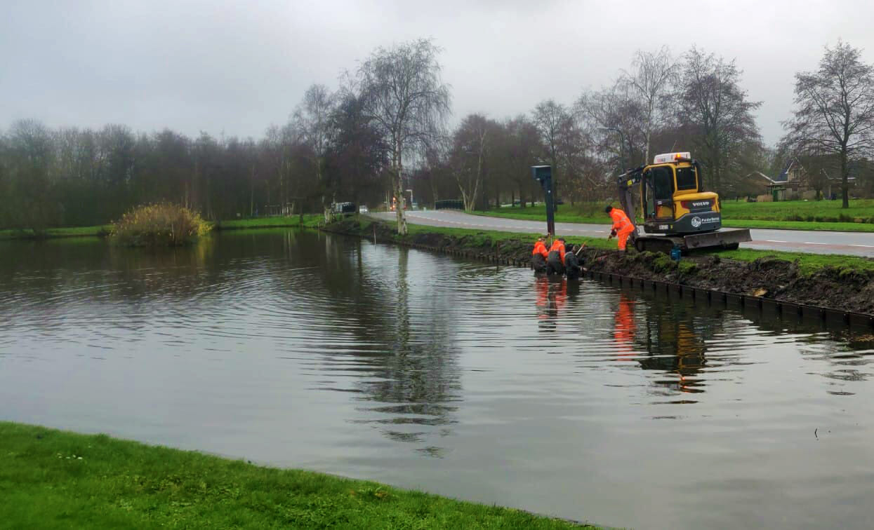 Drie werkers in oranje staan tot aan de knieën in het water van een grote vijver langs een eenbaansweg. Op de oever staat nog een werker en een kleine bobcat graafmachine. Ze zijn de bermwand aan het versterken.